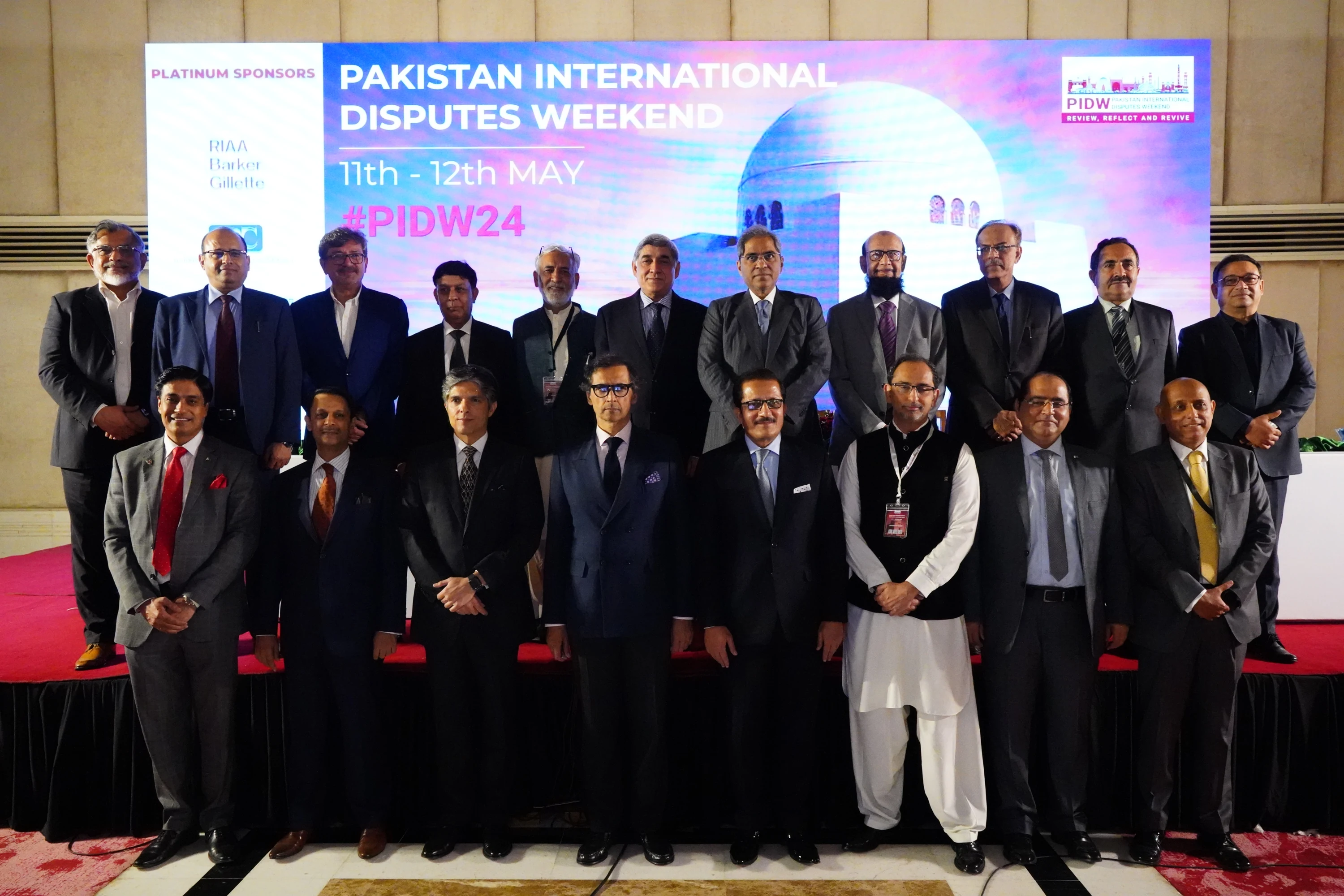 PIDW24 group photograph: eight guests in the front row on the floor and eleven in the back row on the raised stage.